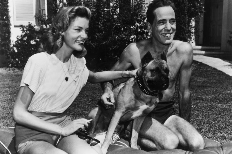 American actor Humphrey Bogart smiles as he kneels with his wife, actor Lauren Bacall, and their pet dog, on a cushion on their front lawn