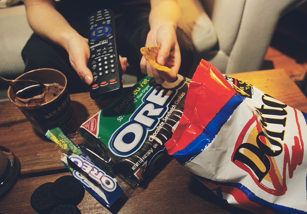 A person holds a remote in one hand and a chip in the other, surrounded by junk food.