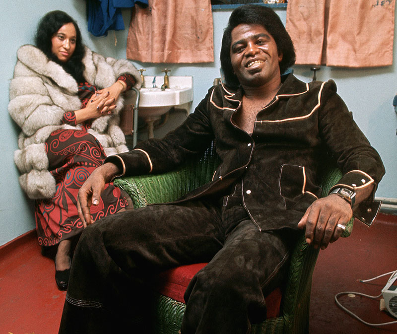 James Brown wears a brown suade suit and leans back in his armchair backstage, smiling to the camera.