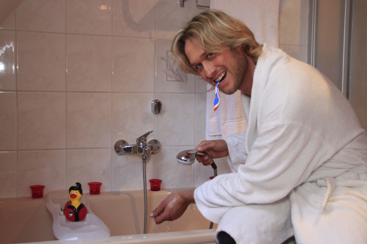 A man kneels near his running bathtub while brushing his teeth.
