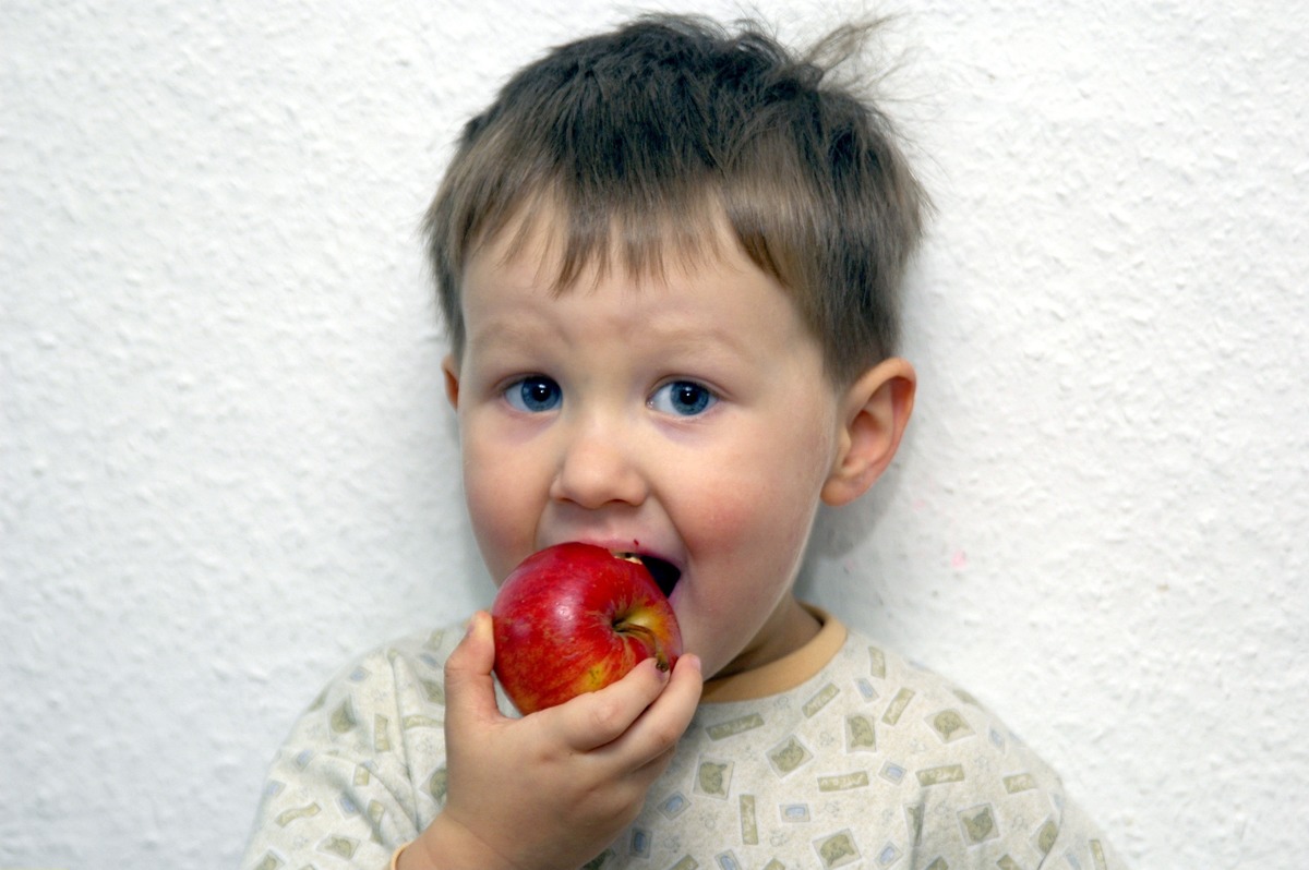 A young boy eats an apple against a white wall.