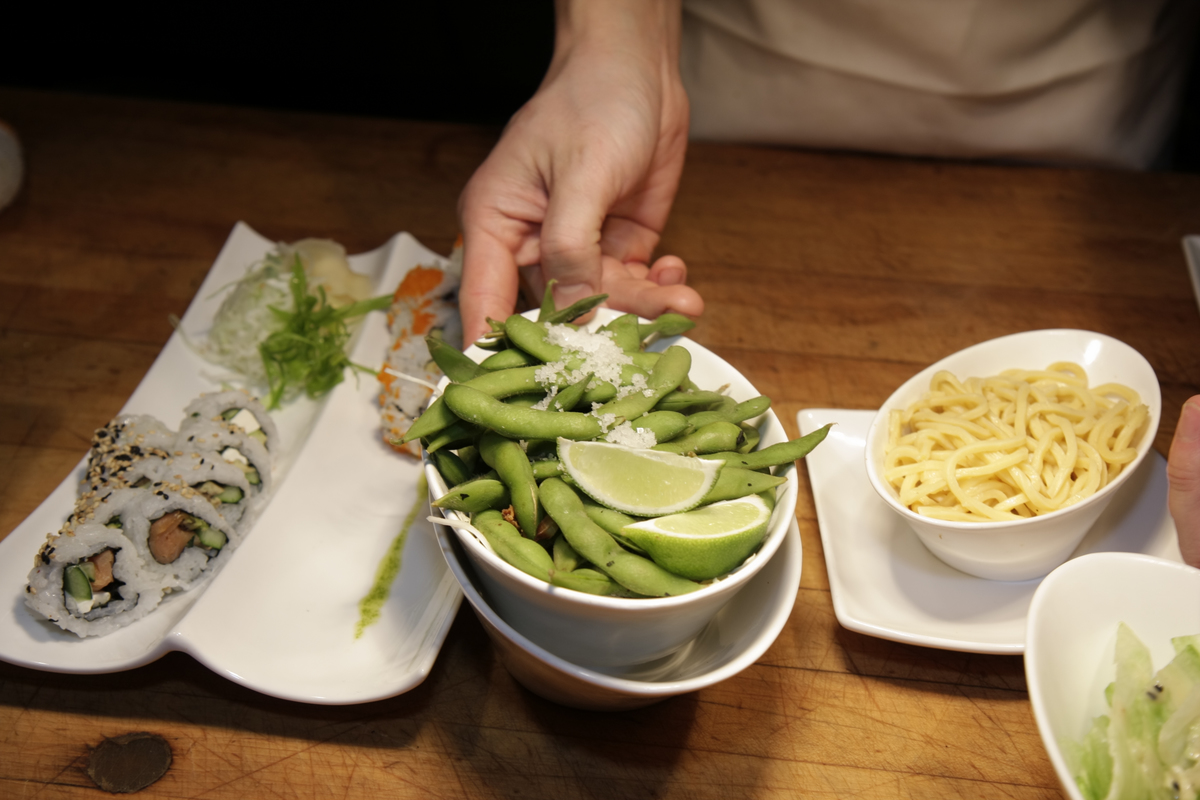 A person holds a bowl of soy over a meal of sushi and low mein.