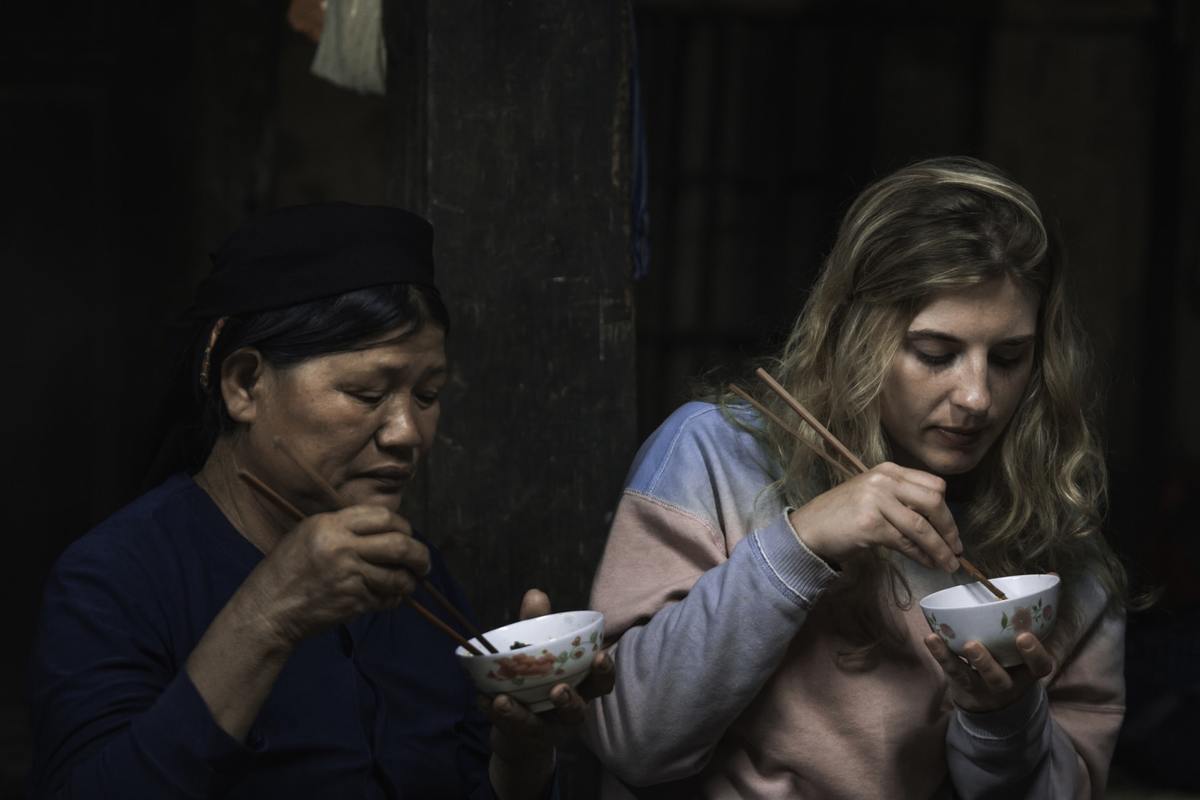 Two women eat from bowls with chopsticks.