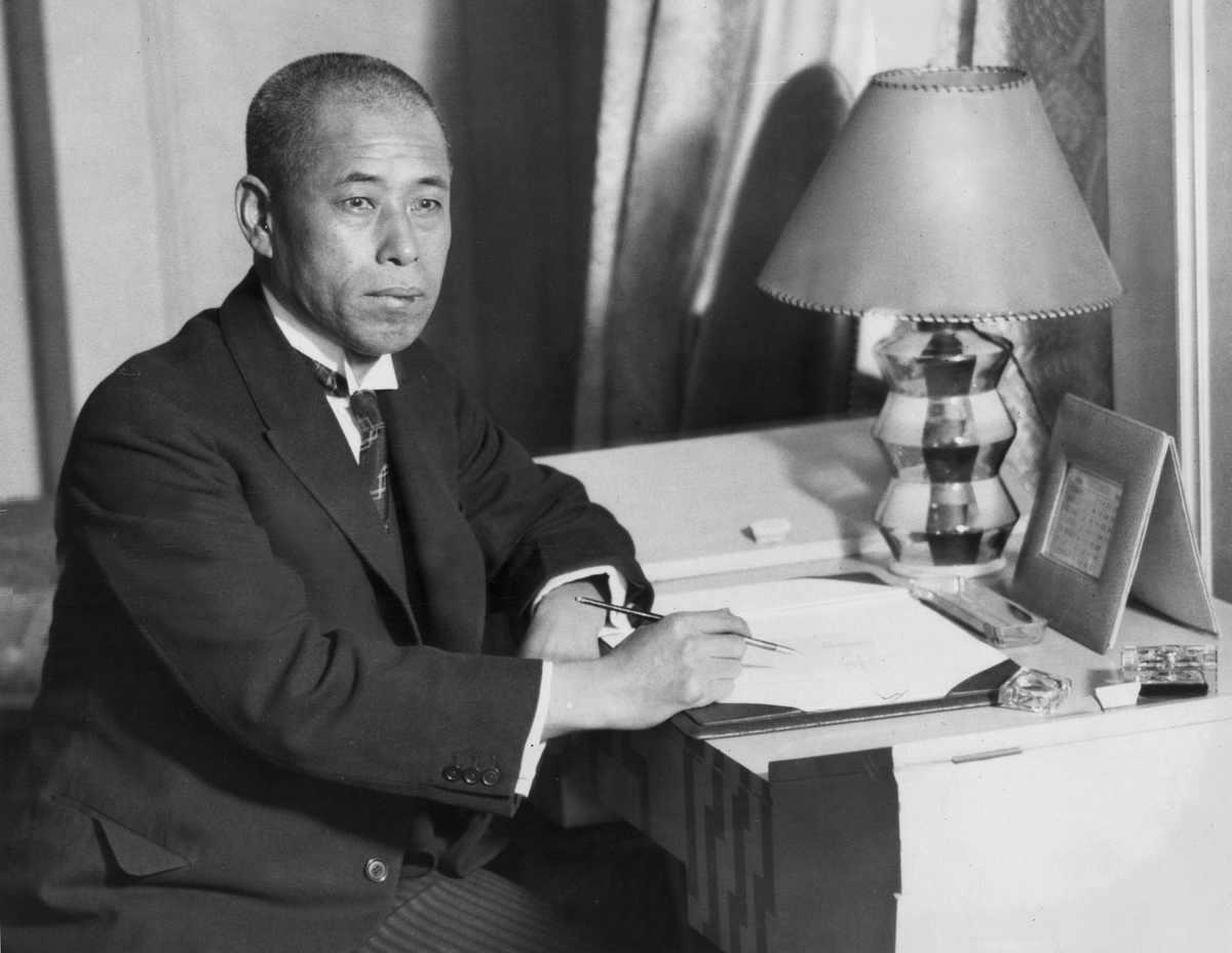 Portrait of Japanese admiral Isoroku Yamamoto, wearing a suit and tie, seated at a desk with a pen in his hand