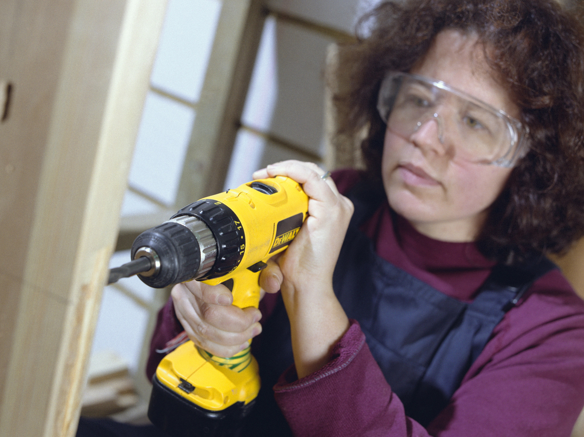 A woman uses a power drill on a wooden plank.