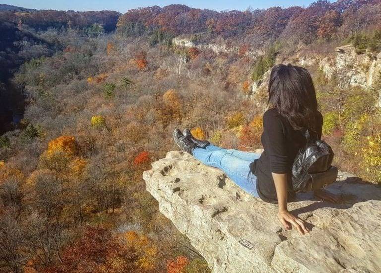 A woman sits on the edge of a rock overlooking a valley of trees.