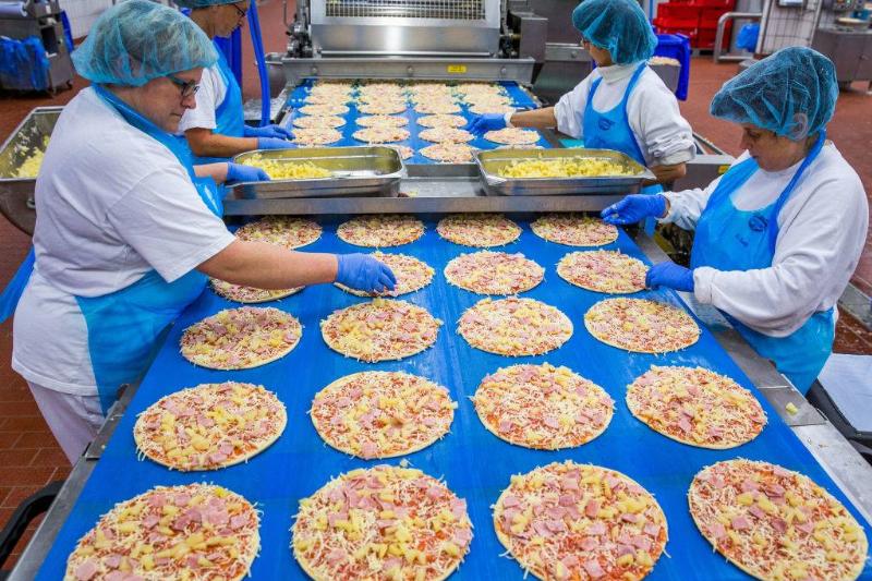 Employees add pineapple pieces to frozen pizzas on a production line