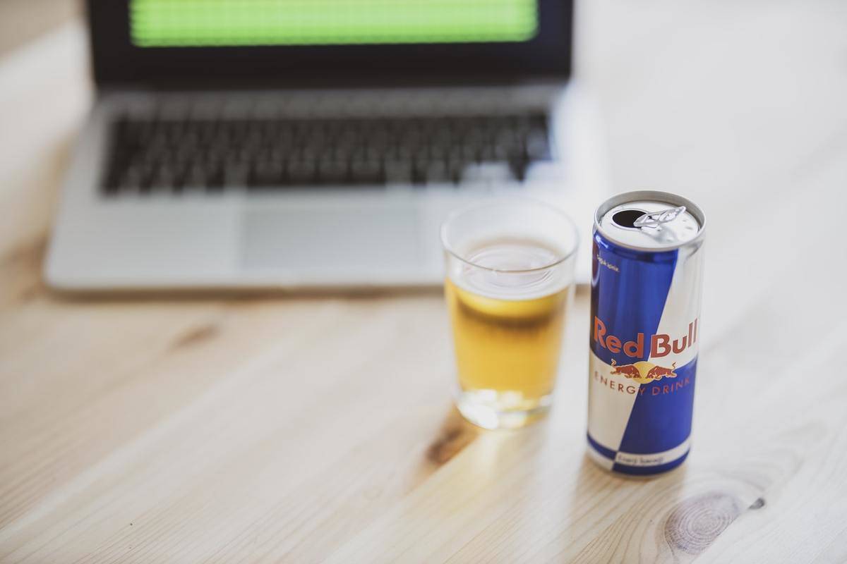 Red bull is poured into a cup next to a laptop.