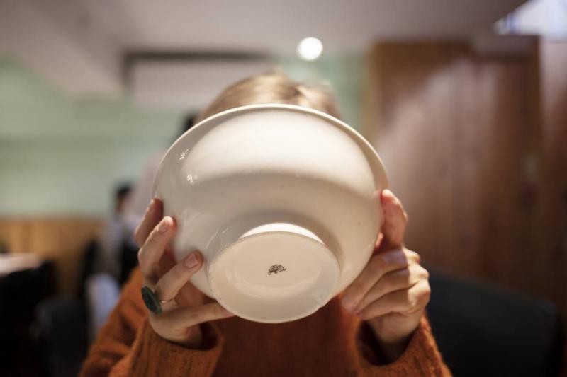 A woman tips a bowl to her face drinking the remainder of her ramen broth.