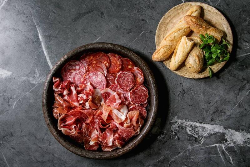 Antipasto meat platter assorti of sliced jamon, salami, chorizo sausage in terracotta tray with bread and greens over black marble background. Flat lay, space