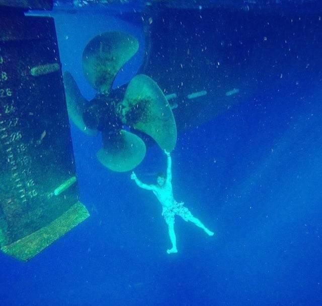 diver posing and doing a thumbs up while holding onto a really big propellor underwater 