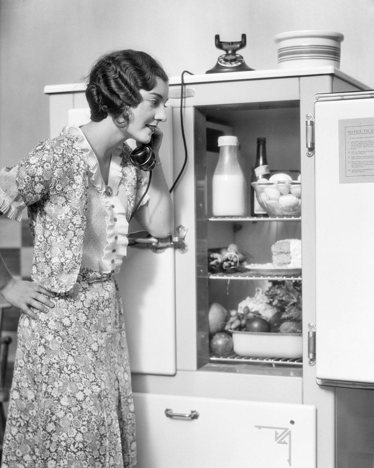 A woman smiles and looks into her refrigerator/icebox while talking on the phone.