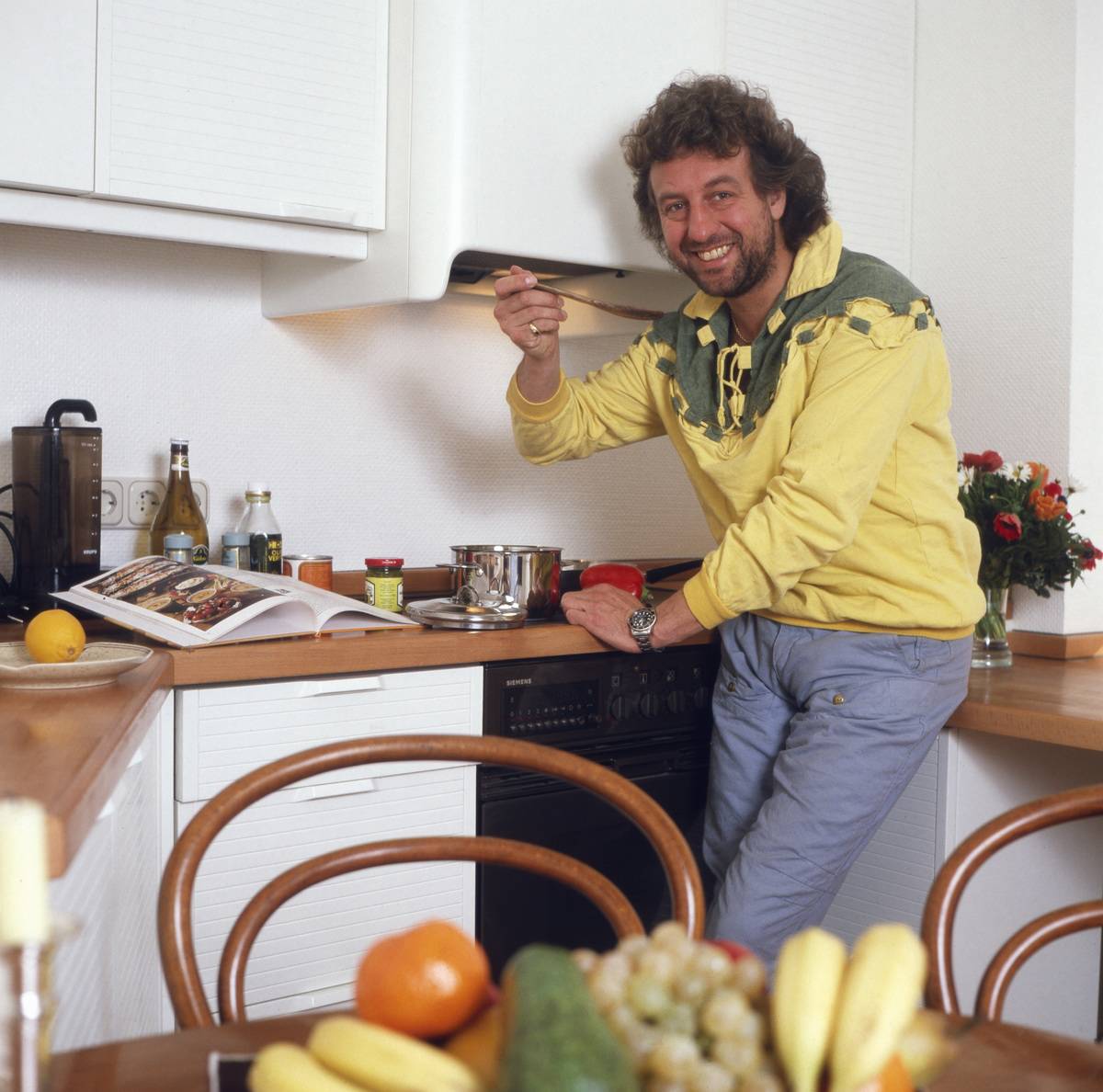 A man tries a sauce that he's cooking in his black and white kitchen, 1980s.