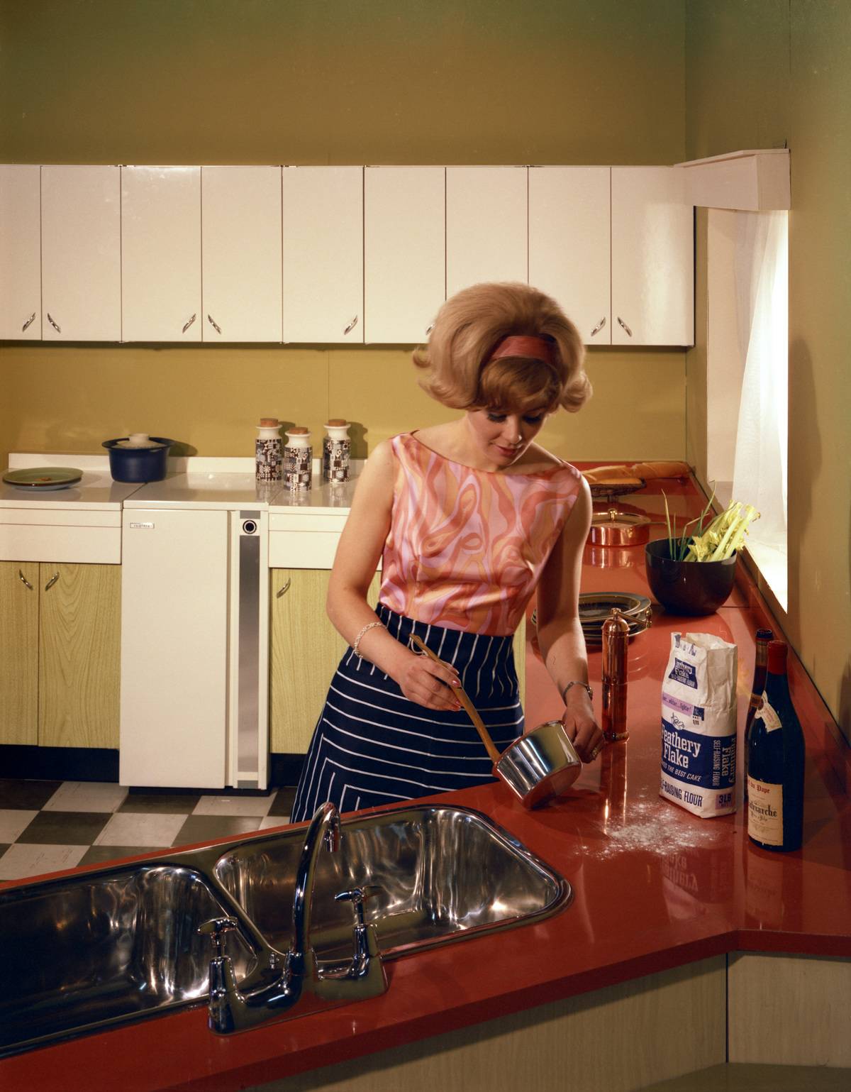 In 1966, a woman cooks in a kitchen with golden walls, a stainless steel sink, and red countertops.