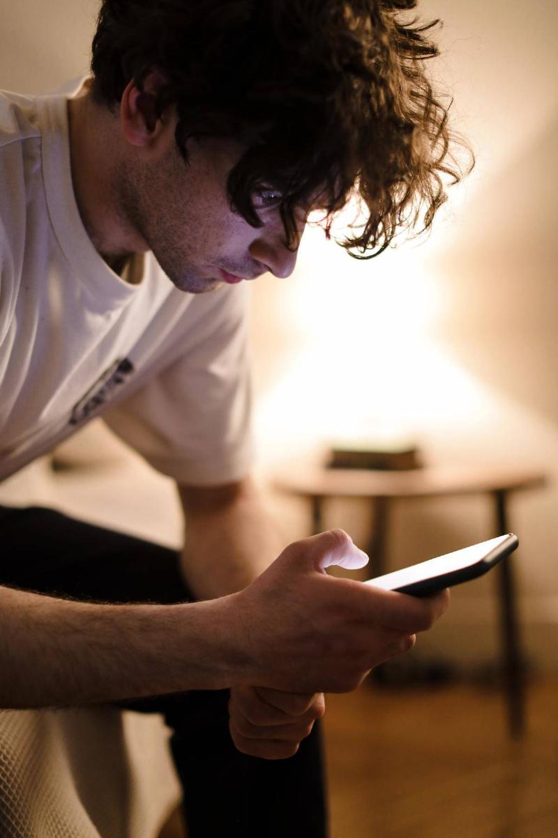 Man seated on edge of bed looking down at cell phone 