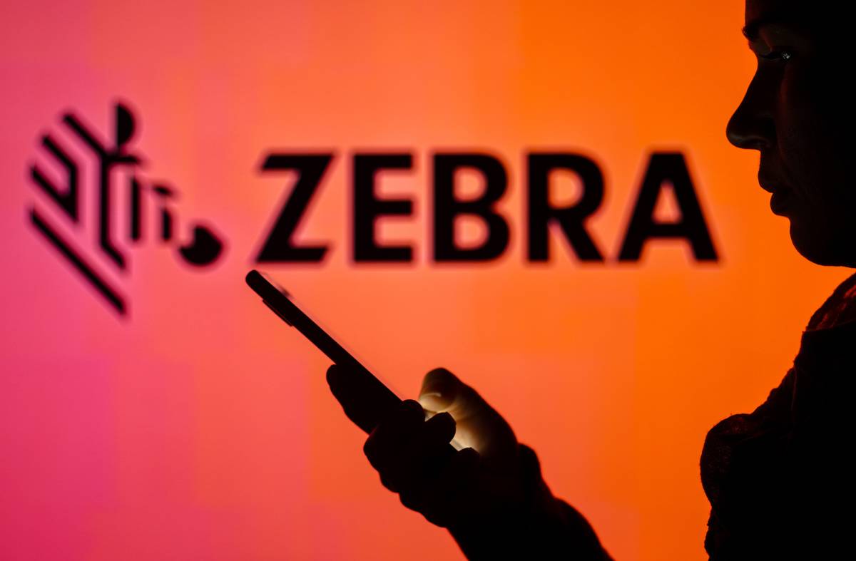 In this photo illustration, the Zebra Technologies logo is seen in the background of a silhouetted woman holding a mobile phone. 