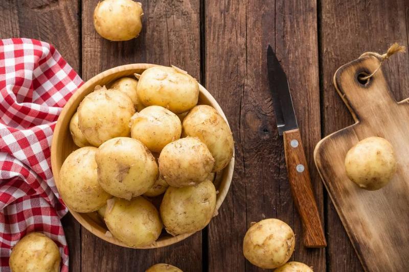 Overhead view of potatoes in a bowl. A wooden cutting board, a knife and a plaid tea-towel are placed nearby
