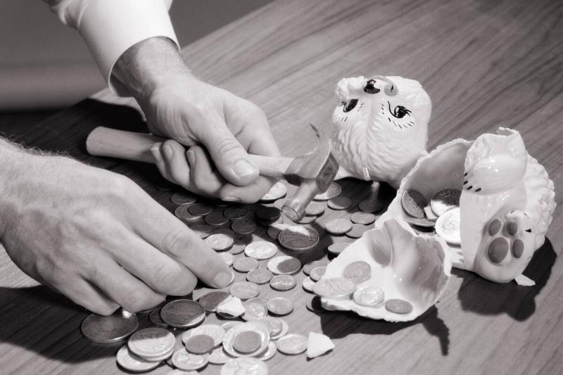 Broken piggy bank and coins scattered across a wooden table. A man's hands cover the scene, with one holding a hammer