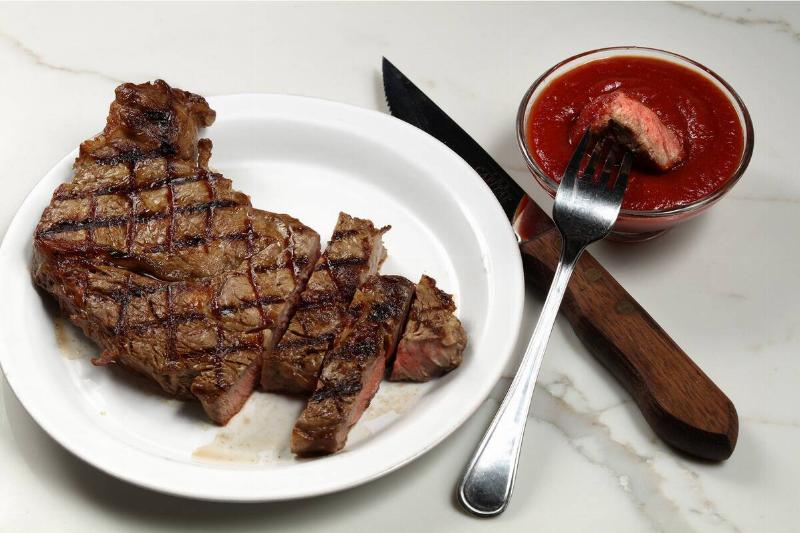 Cut-up steak on a plate, next to utensils and a glass bowl filled with an unidentified red sauce