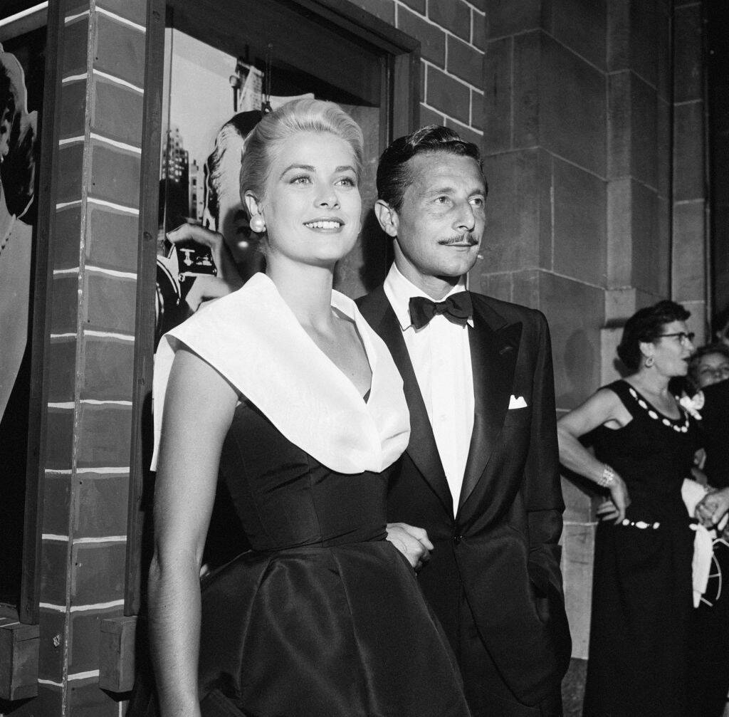 Grace Kelly and Oleg Cassini standing together outside of a building. A group of individuals stand off to the right, slightly behind them