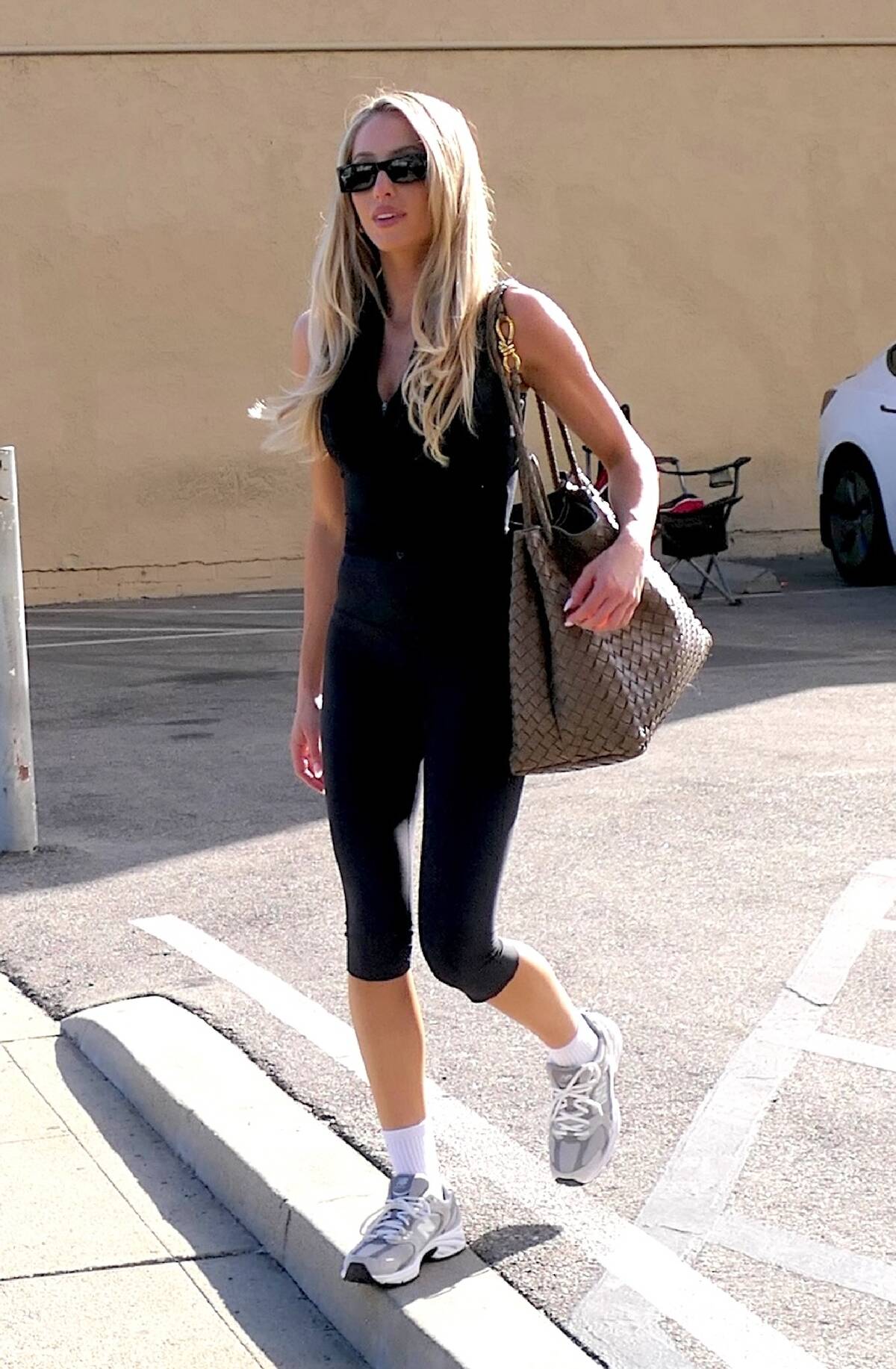 Alix Earle walking into a building from a parking lot. She's dressed in black athletic gear, with a large purse slung over her shoulder