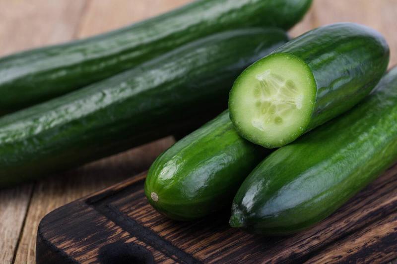 Cut cucumber atop two cucumbers on a wooden cutting board. Two other cucumbers are to the side