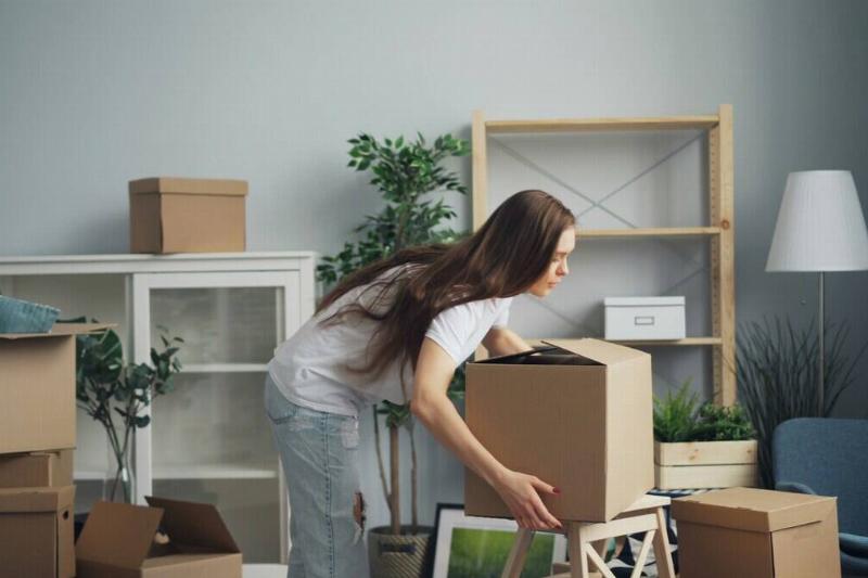 Woman bending over to pick up a cardboard box, in the middle of a room that's half-packed away