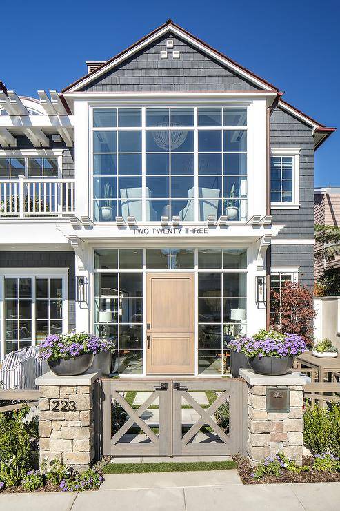Stunning navy blue shingled cottage home boasts a gray gate opening to a backyard fitted with a walnut stained back door framed by glass panels lit by modern iron and glass sconces fixed beneath a second floor pergola.