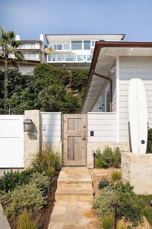 Rustic stone pavers lead to a salvaged wood gate accenting a light gray shingled beach cottage home.